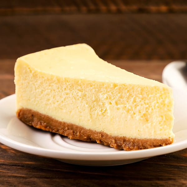 A slice of classic cheesecake with a graham cracker crust on a white plate.