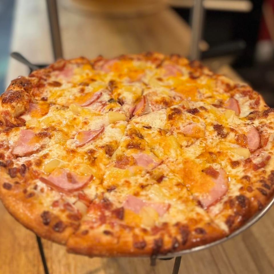 A whole Hawaiian pizza with ham and pineapple on a metal stand.